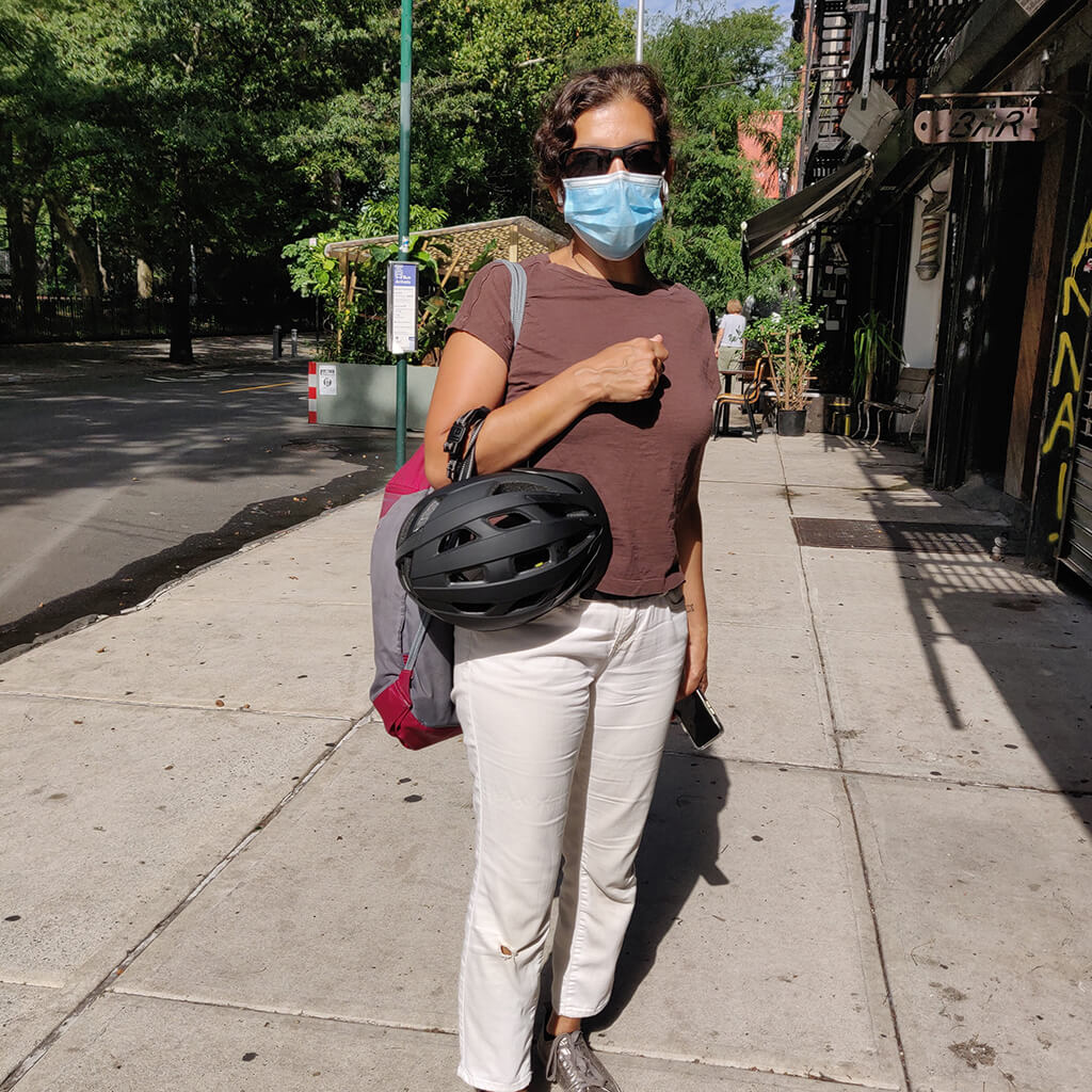 Masked lady standing on sidewalk in morning sunshine with helmet on her arm